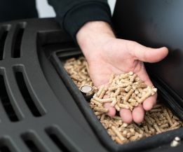 Eine Hand hält Holzpellets über dem geöffneten Brennraum einer Pelletheizung, Symbol für erneuerbare Heizenergie